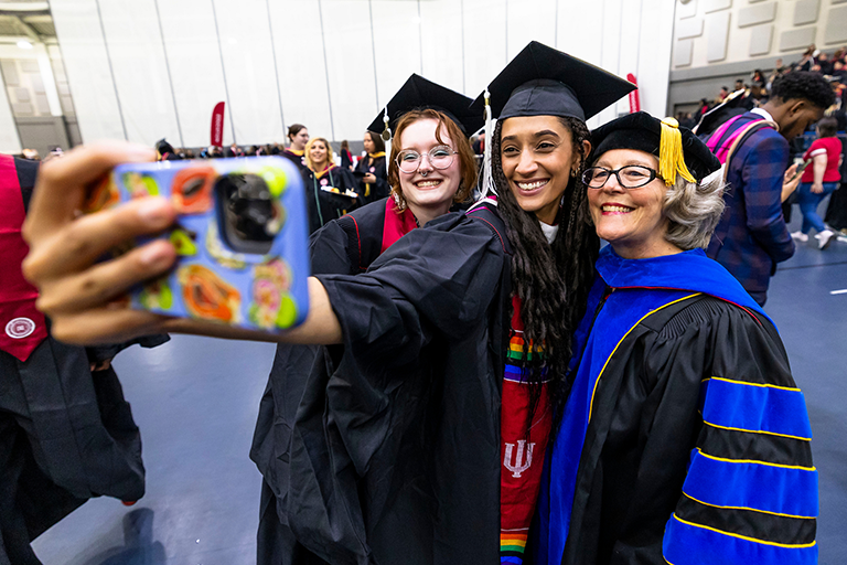 Two female masters students and a woman professor pose in front of a phone camera to take a selfie at graduation.